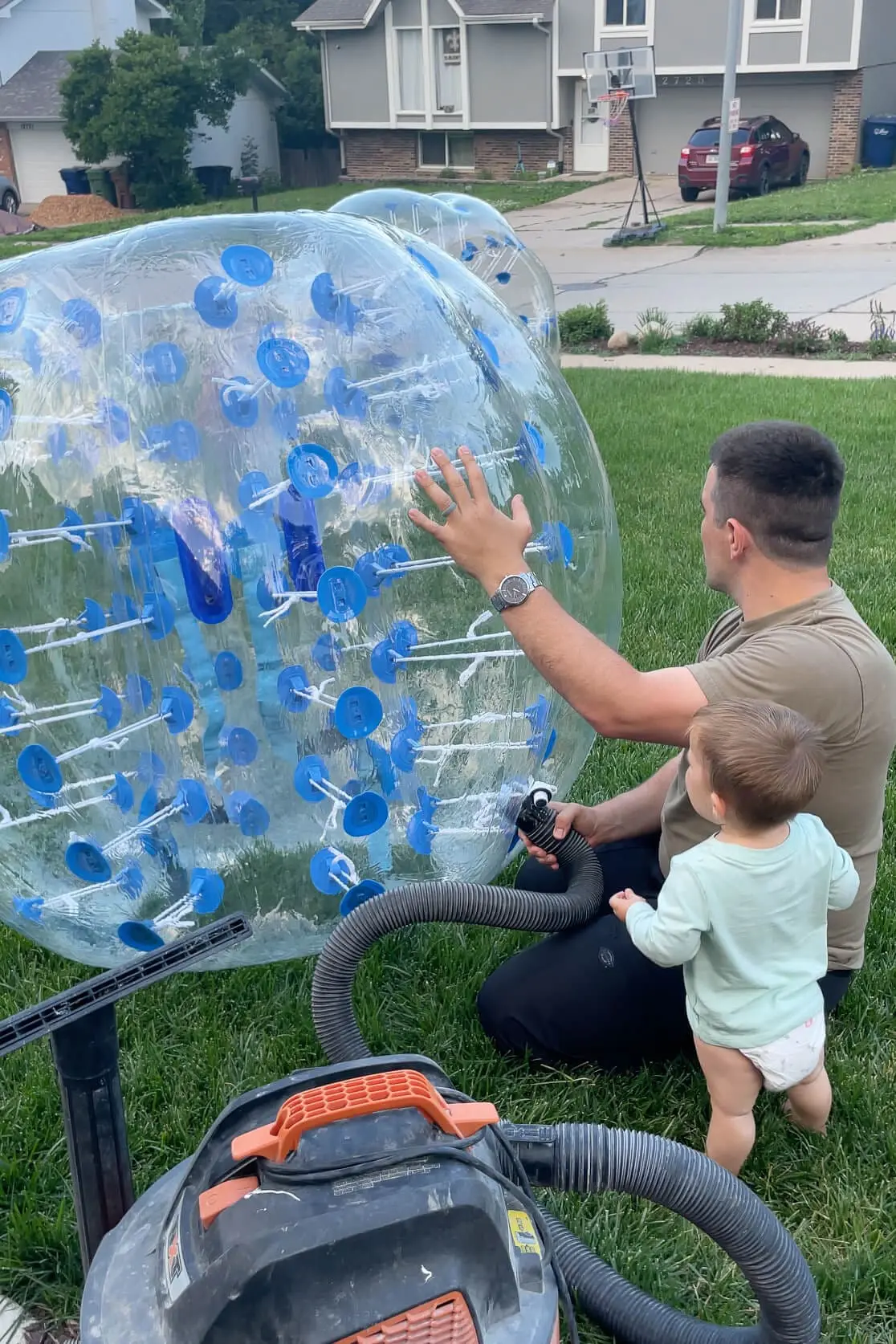 Father and toddler using a vacuum to inflate a large bumper ball, prepping for a day of fun backyard activities.